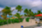 Beach scene with palm trees and colorful kayaks. Empty lounge chairs line the sandy shore. A "Watersport Rentals" hut with a red roof is visible.