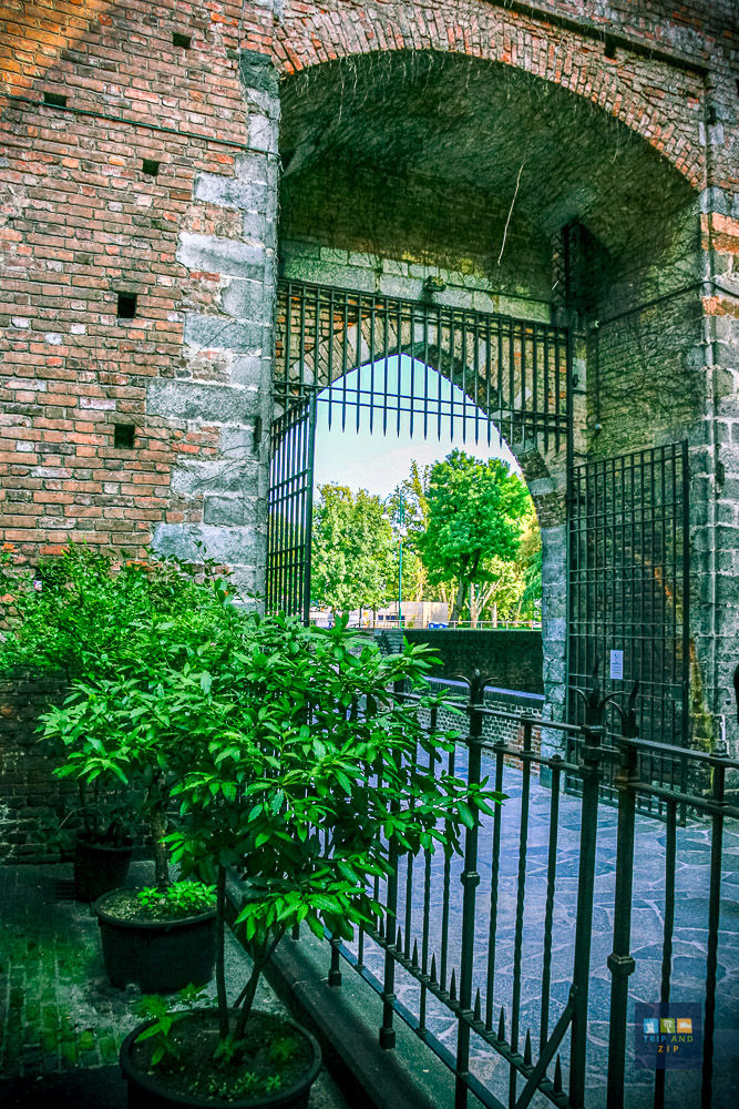 Old brick archway with iron gate, potted green plants, and vibrant foliage in background. Cobblestone path suggests a historic setting.