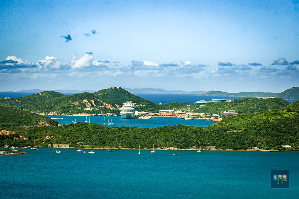 Cruise ship docked in a turquoise bay, surrounded by lush green hills under a bright sky with scattered clouds. Calm and scenic.
