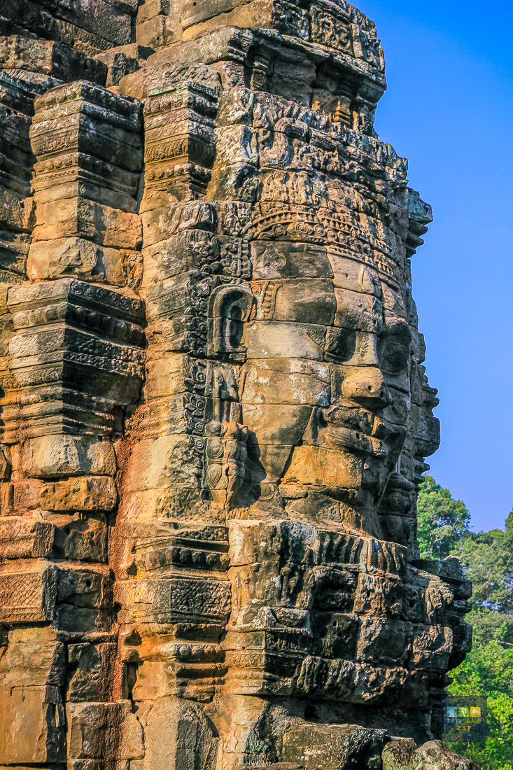 Stone face sculpture on an ancient temple wall, detailed carvings, warm hues, vivid blue sky, green trees in background. Calm and majestic.