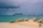 Cruise ship on blue sea under cloudy sky, with small boats and kayakers nearby. Sandy beach with rocks, umbrella, and a person sitting.