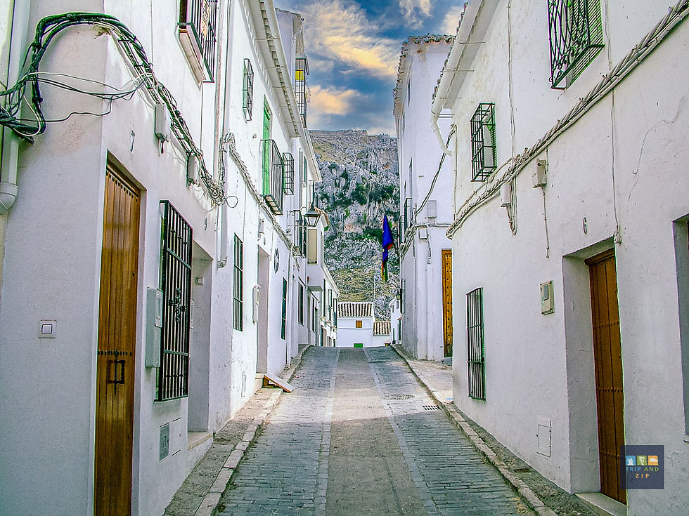 Narrow cobblestone street with white buildings, green windows, and flags. Mountain view under a cloudy blue sky. Quiet, scenic atmosphere.