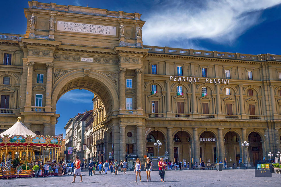 People walk near an ornate arch in a sunny plaza, with a colorful carousel on the left. The building reads Pensone Pendini.
