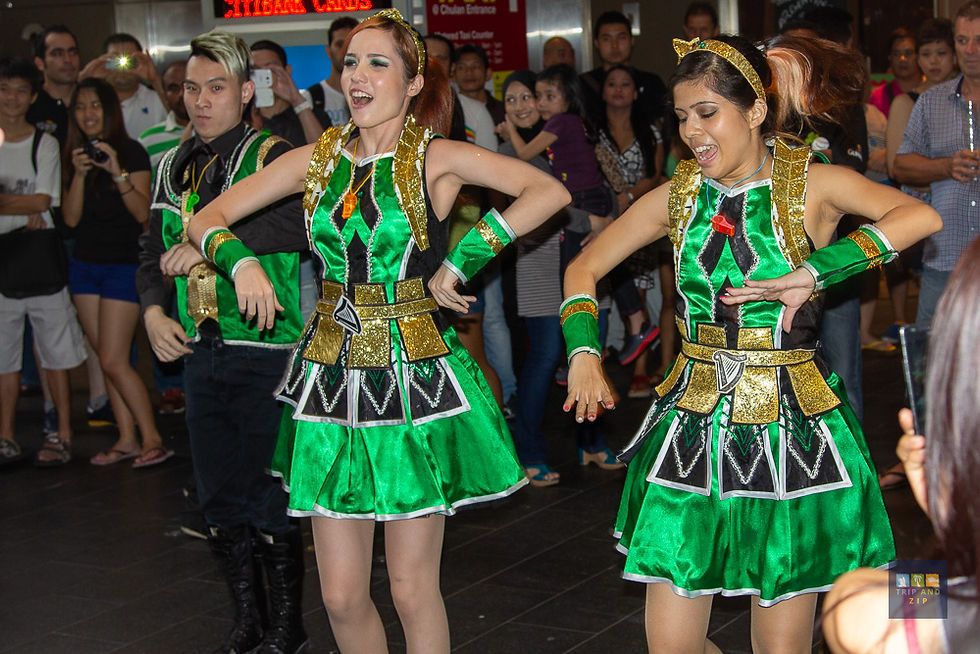 Performers in green and gold costumes dance energetically in a crowded indoor space. Onlookers watch, creating an upbeat and lively mood.