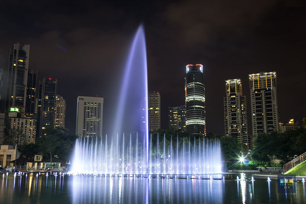 Nighttime cityscape with a tall, illuminated water fountain against a backdrop of lit skyscrapers in Kuala Lumpur. Reflections on water.