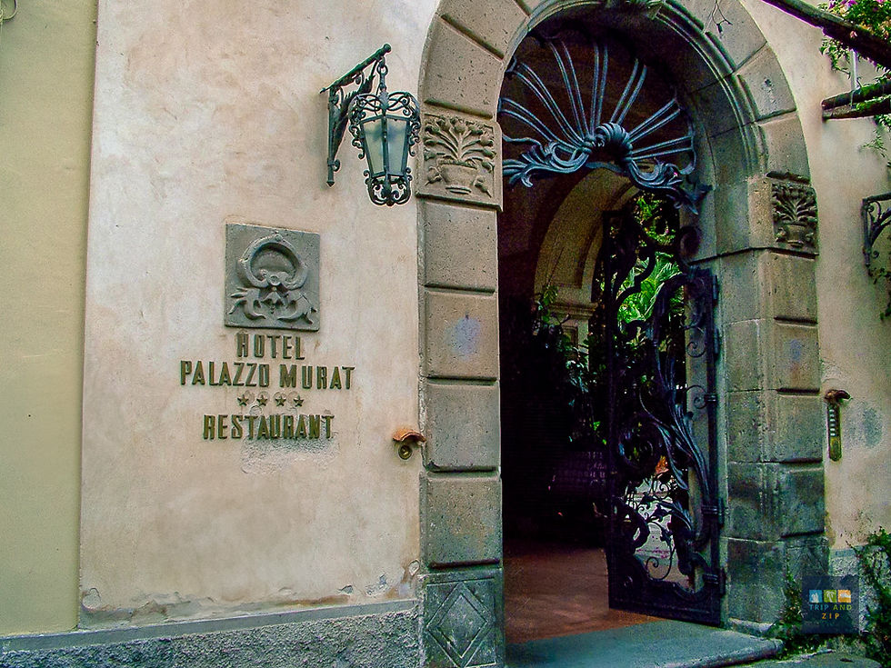 Stone archway with ornate black wrought-iron gate, surrounded by greenery. Wall reads "Hotel Palazzo Murat Restaurant" with star symbols.
