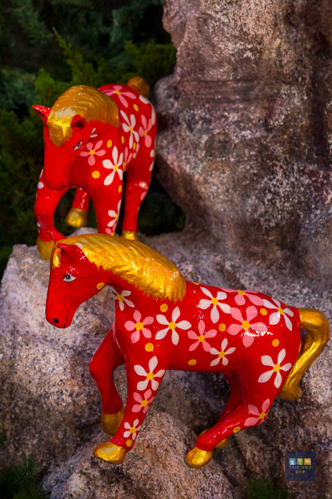 Two red, floral-patterned horse sculptures with gold manes set against a rocky background and greenery, creating a vibrant scene.