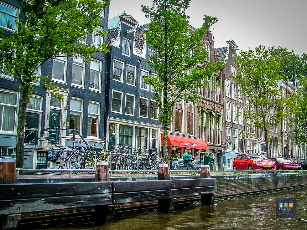 Canal view of Amsterdam buildings, parked bikes, red cars. Trees line the street. A person sits by the "red appels" cafe. Moody ambiance.