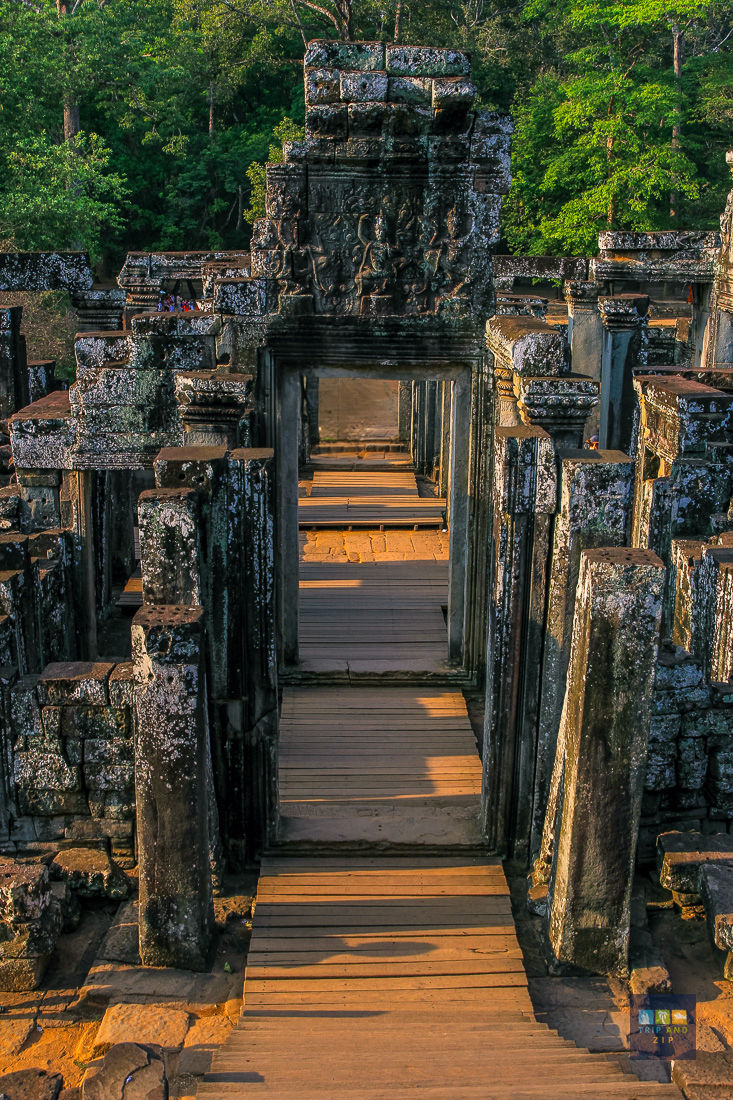Ancient stone ruins with intricate carvings, surrounded by lush greenery. Warm sunlight casts shadows on the wooden pathway through arches.