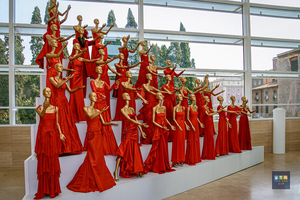 Mannequins in red dresses are posed dramatically on tiered stands in a glass-walled room, with greenery visible outside.