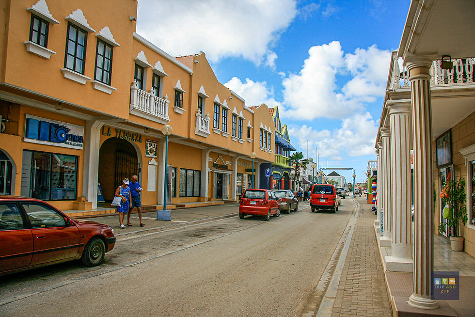 Street scene with colorful buildings, cars parked alongside. Two people walk, one carries a bag. Blue sky with clouds, relaxed atmosphere.