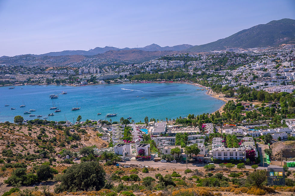 Coastal cityscape with turquoise bay, boats, and a mountainous backdrop. White buildings and greenery surround the vibrant shoreline.