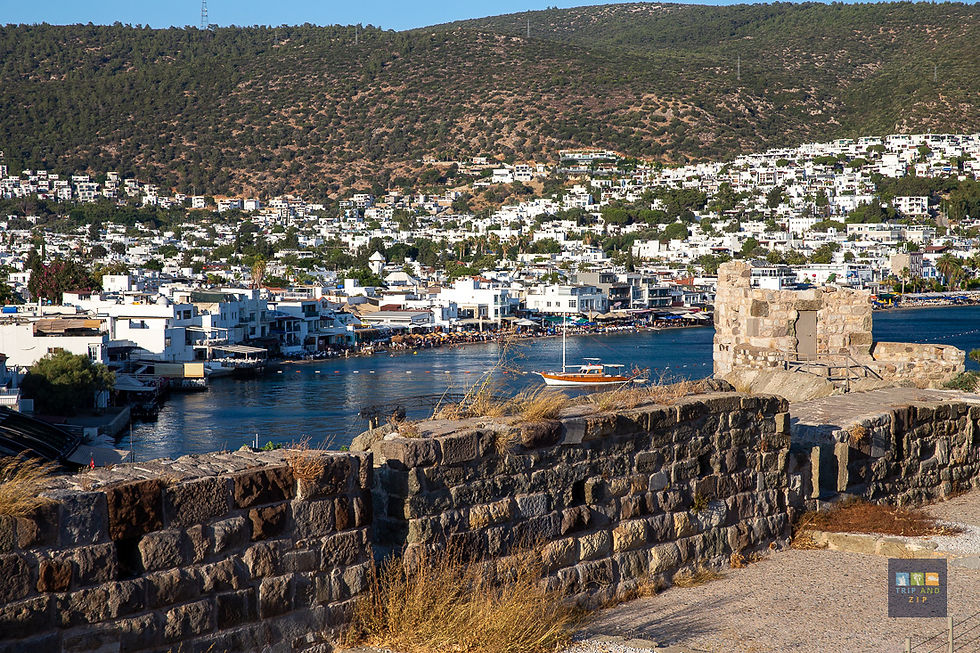 View of a coastal town with white buildings, a hill backdrop, and a stone wall in the foreground. A wooden boat is on the blue water.