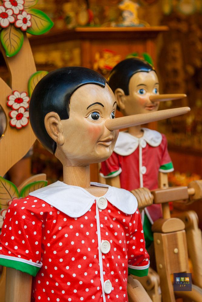 Wooden Pinocchio figures with long noses in red polka-dot outfits. Background includes colorful floral decorations and shelves.