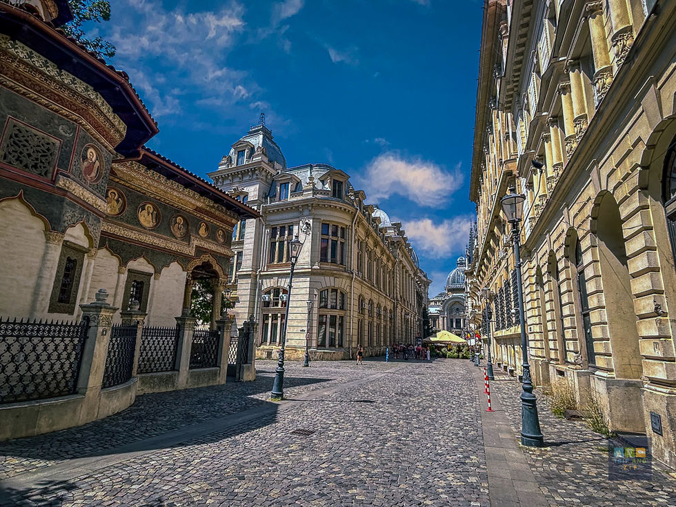 Historic street scene with ornate buildings, cobblestone path, and vibrant blue sky. People walk near outdoor café with yellow umbrella.