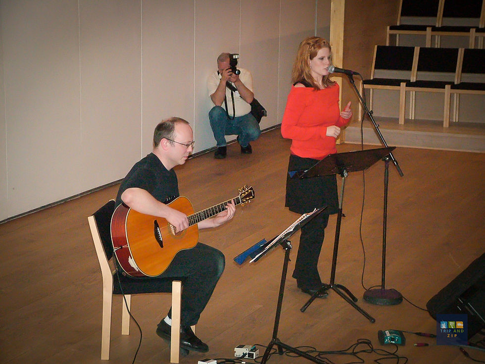 A woman sings into a microphone, wearing a red top. A man beside her plays a guitar. A photographer kneels in the background.