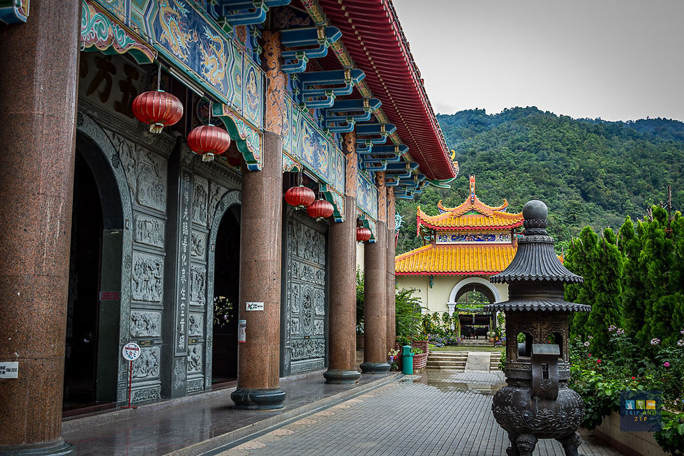 Colorful temple with ornate columns and red lanterns against a mountainous backdrop. Stone path leads to an intricately designed incense burner.