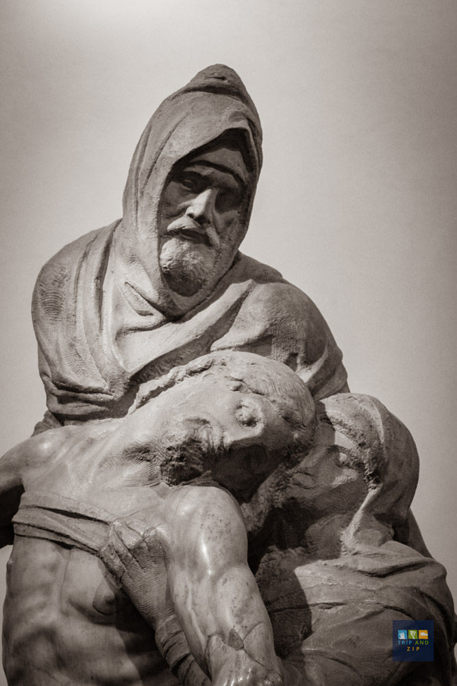 Stone sculpture of a hooded figure supporting a limp body, evoking somber emotion. Neutral background, small logo at bottom right.