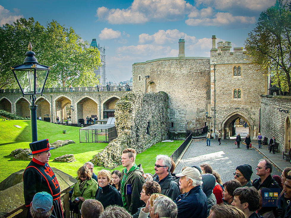 Tour guide in black and red uniform leading a group outside a historic stone castle under a blue sky. Green lawns and trees surround.