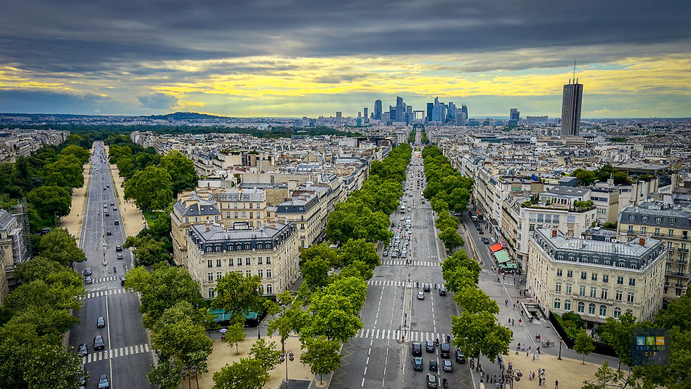 Aerial view of Paris streets with green trees, leading to La Défense skyline under a cloudy sky. Bustling street life with vehicles.