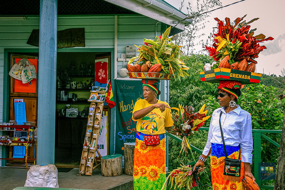 Women in vibrant skirts balance baskets of produce on their heads near a shop. Signs read "Grenada." Lush greenery surrounds them.