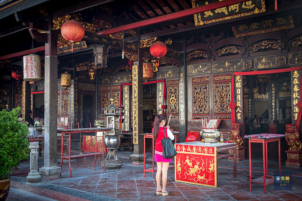 Woman in red stands in prayer at ornate temple entrance with red lanterns and gold details. Incense smoke rises; mood is serene.