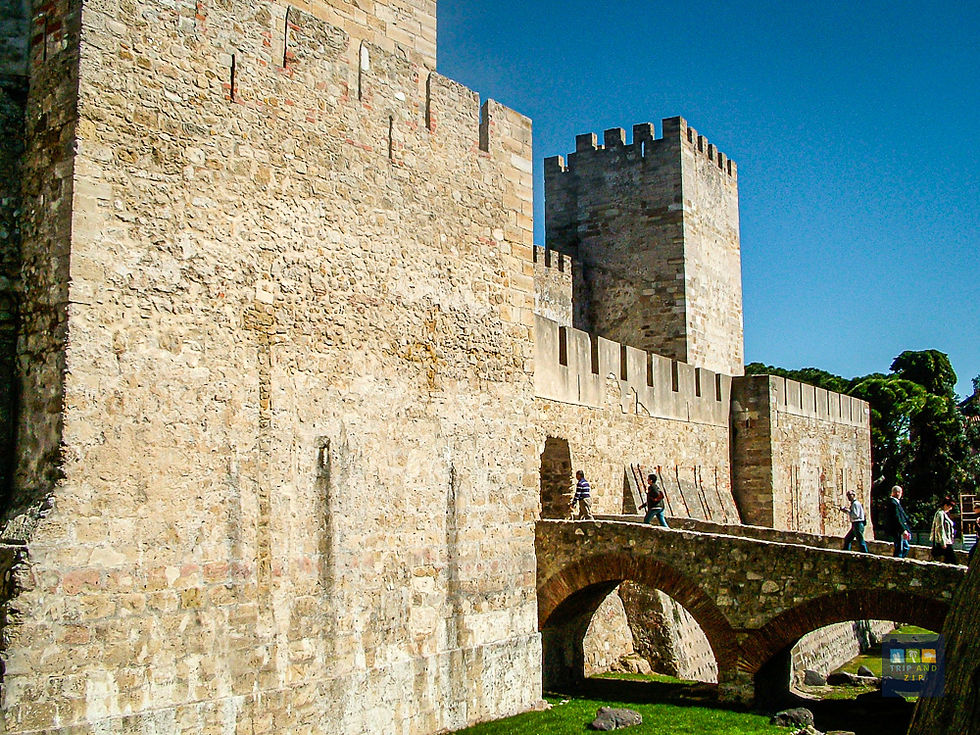 Stone castle with tall towers in bright daylight. People walk on an arched bridge. Blue sky and greenery in the background.