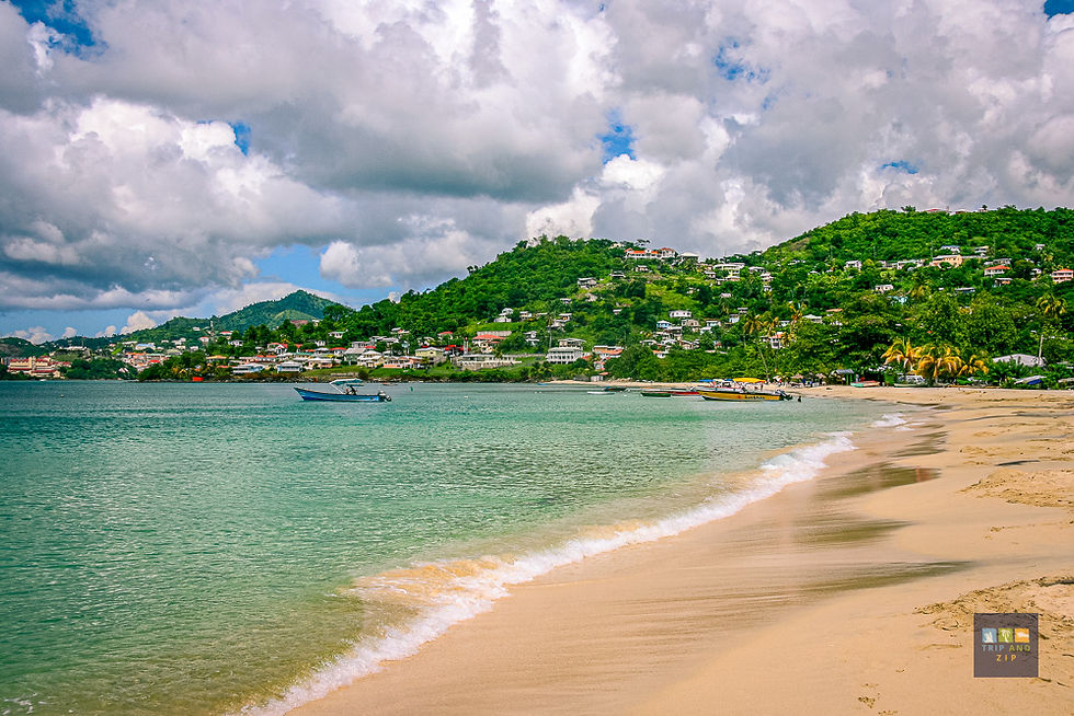 Sandy beach with turquoise water and small boats. Lush, green hills with houses in the background under cloudy skies. Mood is tranquil.