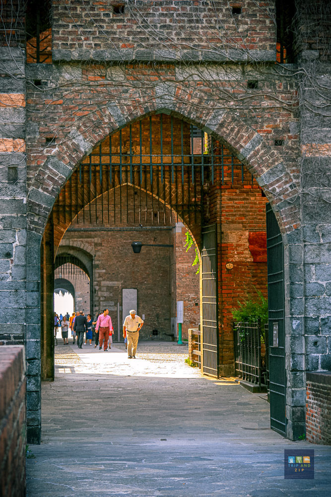 Stone archway at a castle with people walking beneath. Sunlight illuminates the path. Green vines partially cover the red brick.