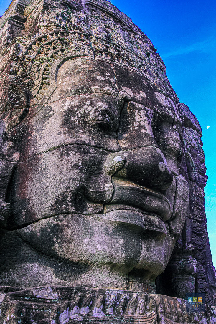 Close-up of a stone face sculpture with intricate carvings under a clear blue sky. The moon is visible in the background.