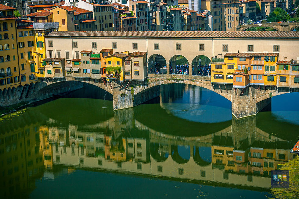 Historic bridge with yellow buildings over a calm river, reflecting the architecture. Crowds visible. Cityscape in the background.