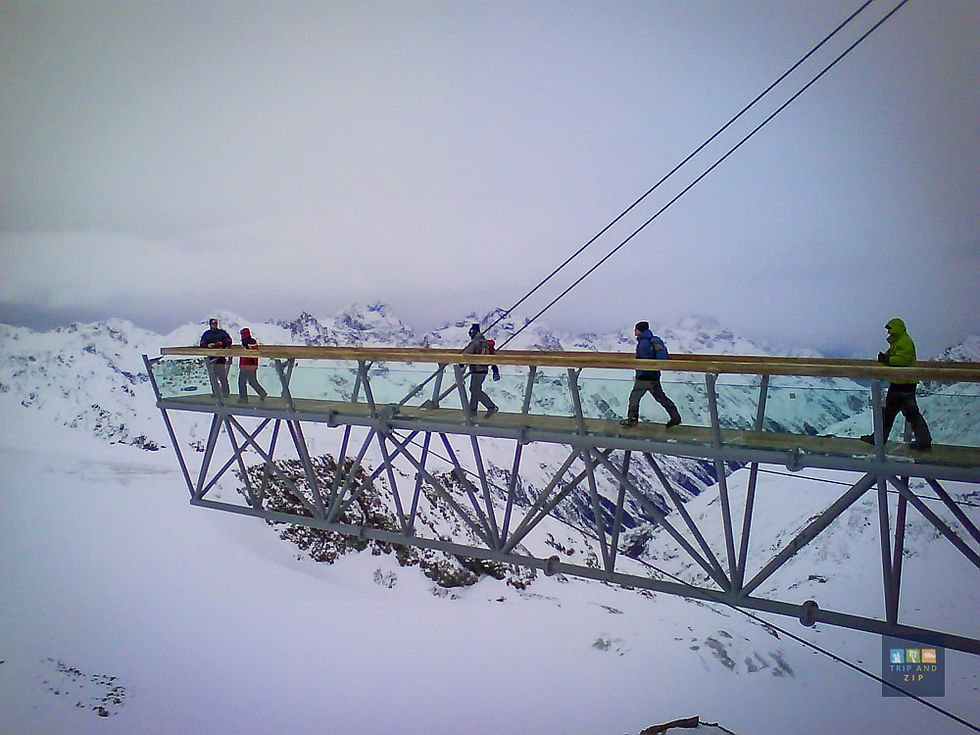 People in winter clothing walk across a glass bridge over snowy mountains. The sky is overcast, creating a serene and adventurous mood.