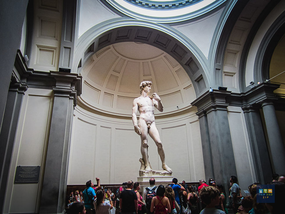 People admire a large marble statue in a grand hall with arched ceilings. A plaque is visible on the left. Mood is awe-inspired.