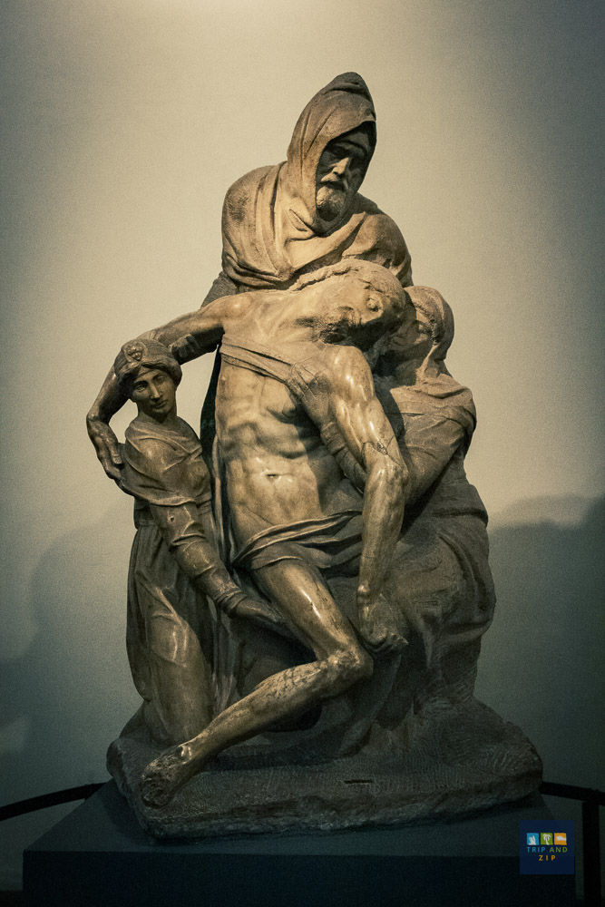 A somber marble sculpture depicts three figures supporting a lifeless body. The background is plain; colors are muted. Text reads "Trip and Zip."