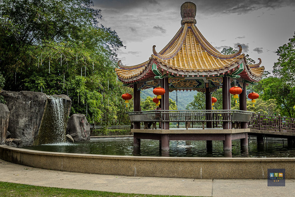 Chinese pagoda with red lanterns over a pond, surrounded by lush greenery and a waterfall. The sky is overcast, creating a serene mood.