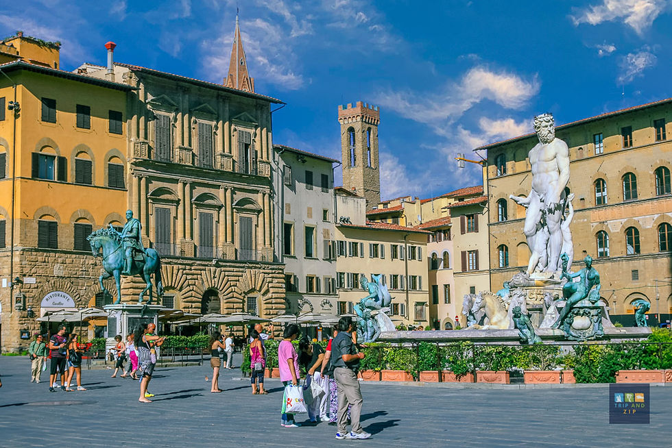 Tourists walk in a sunny plaza with historic buildings, a fountain with statues, and a turquoise equestrian monument. Bright and lively setting.
