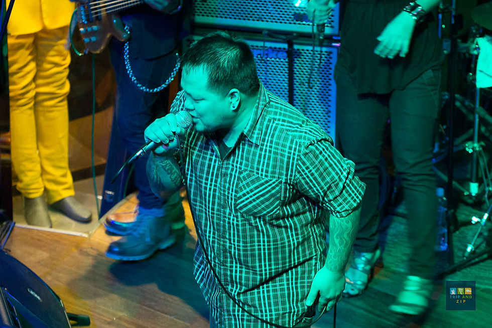 Performer in plaid shirt singing into mic on stage, surrounded by musicians with colorful outfits; lively atmosphere.