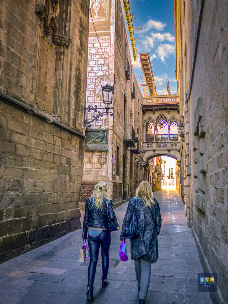 Two women in jackets walk through a narrow, historical stone street under a blue sky. They carry shopping bags, creating a leisurely vibe.