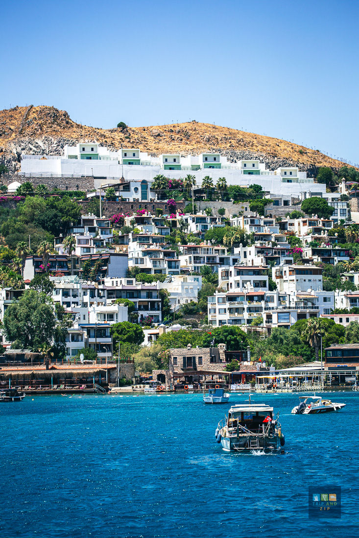 Boats on clear blue water with a hillside backdrop of colorful houses and greenery under a bright blue sky. Relaxing coastal scene.