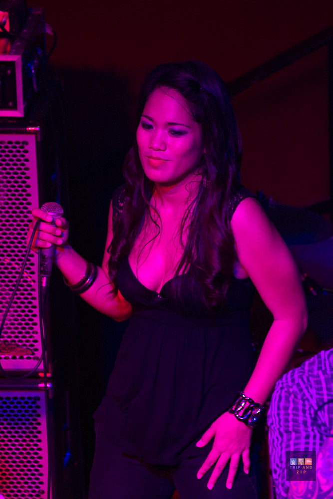 Singer holding microphone on stage, wearing black top under purple light. Amplifiers in background. Focused expression. Text: "TRIPANDZIP".