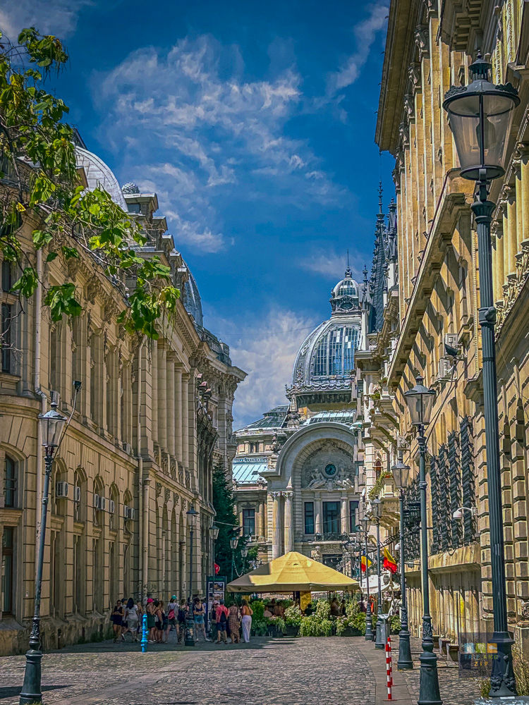 Street view with historic buildings and a central, dome-topped structure. People gather near a yellow umbrella. Bright, sunny day.
