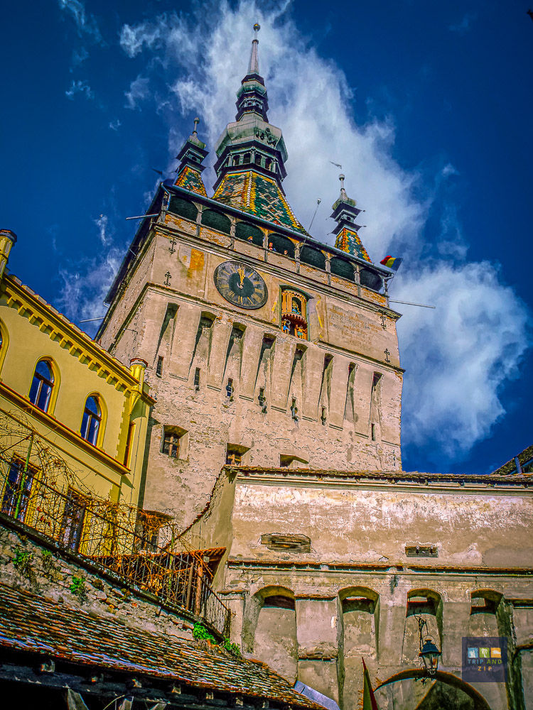 Tall medieval clock tower with pointed roofs and vibrant tiles under a partly cloudy blue sky. Romanian flag waves, creating a historical ambiance.