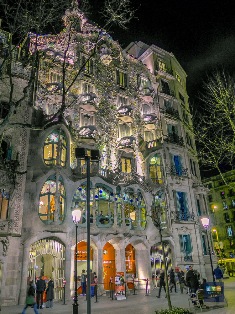 Illuminated night view of Casa Batlló in Barcelona with colorful mosaic facade, people walking, and leafless trees in the foreground.