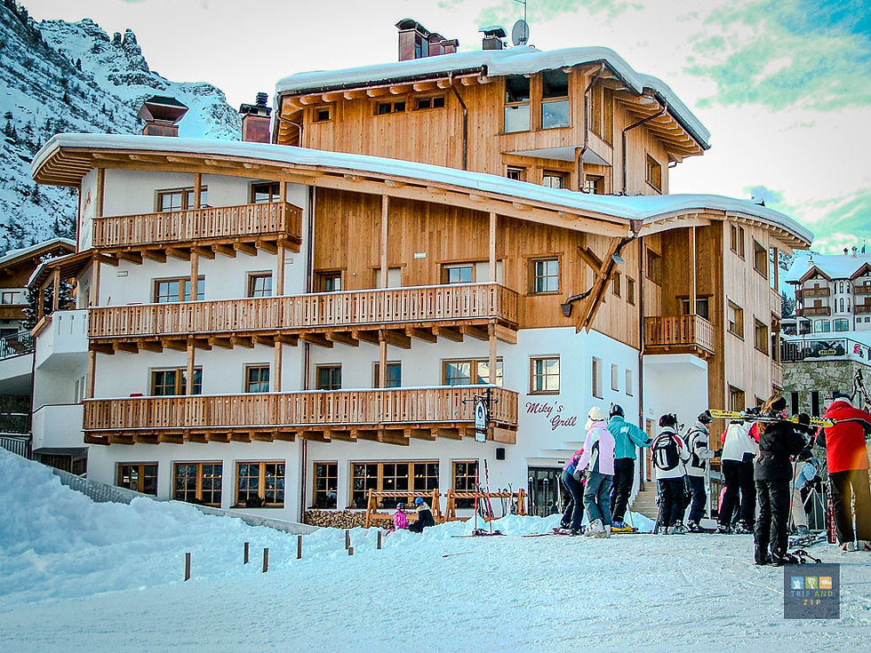 Skiers gather outside a wood-accented alpine lodge named "Miky's Grill" in a snowy mountain setting. Bright winter clothing contrasts with snow.