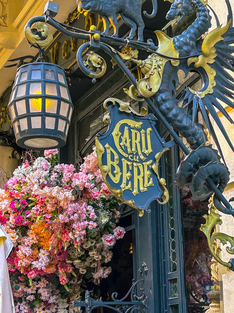 Ornate sign reading "Caru cu Bere" hangs amid colorful flowers outside a building. Features a dragon, lantern, and intricate metalwork.