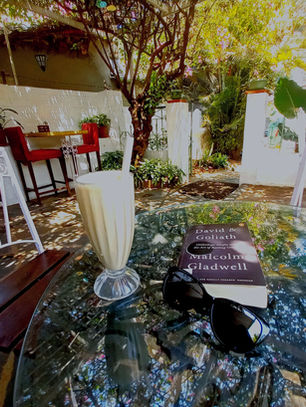 In a cafe nestled amidst trees and plants, on a glass table lies a frappe, a pair of black sunglasses and a copy of Malcolm Gladwell's David and Goliath. Photo by Ninay Desai.