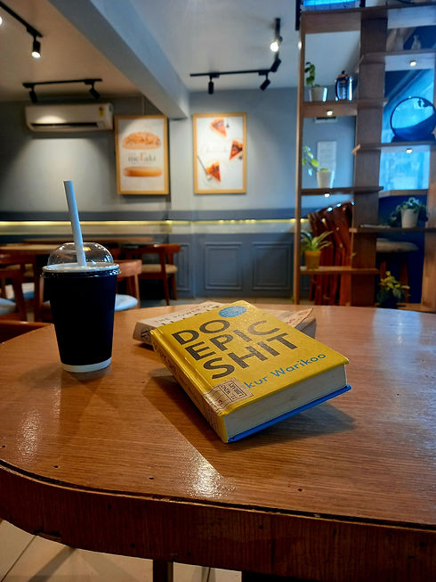 In a cafe, a copy of Do Epic Shit by Ankur Warikoo lies on a round, wooden table next to a frappe in a disposable cup. Photo by Ninay Desai.
