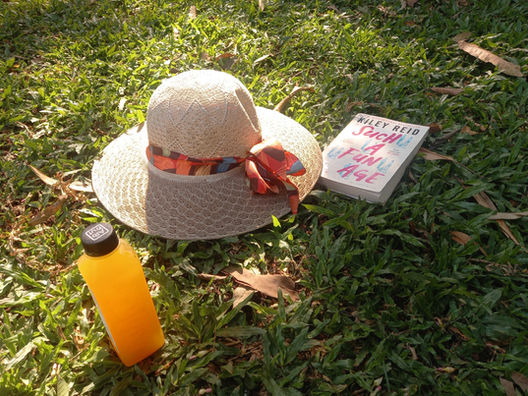 A copy of Kiley Reid's Such a Fun Age is lies in the grass. A straw hat with a broad multi-coloured ribbon is tossed close to the book. Next to it lies a small bottle with an orange drink. Photo by Ninay Desai.