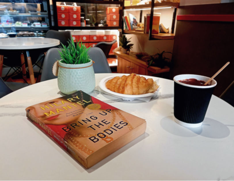 On a white round table in a cafe, lies a copy of Hilary Mantel's historical fiction novel Bring up the Bodies. Beside it are a cup of hot chocolate, a croissant and a small plant. In the background are shelves displaying the cafe's goods. Photo by Ninay Desai
