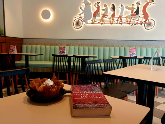 On a white table next to an ice cream sundae lies Chitra Banerjee Divakaruni's novel, Sister of My Heart. In the background are a few more tables and chairs, and an illuminated wall art on a cream-coloured wall. Photo by Ninay Desai.
